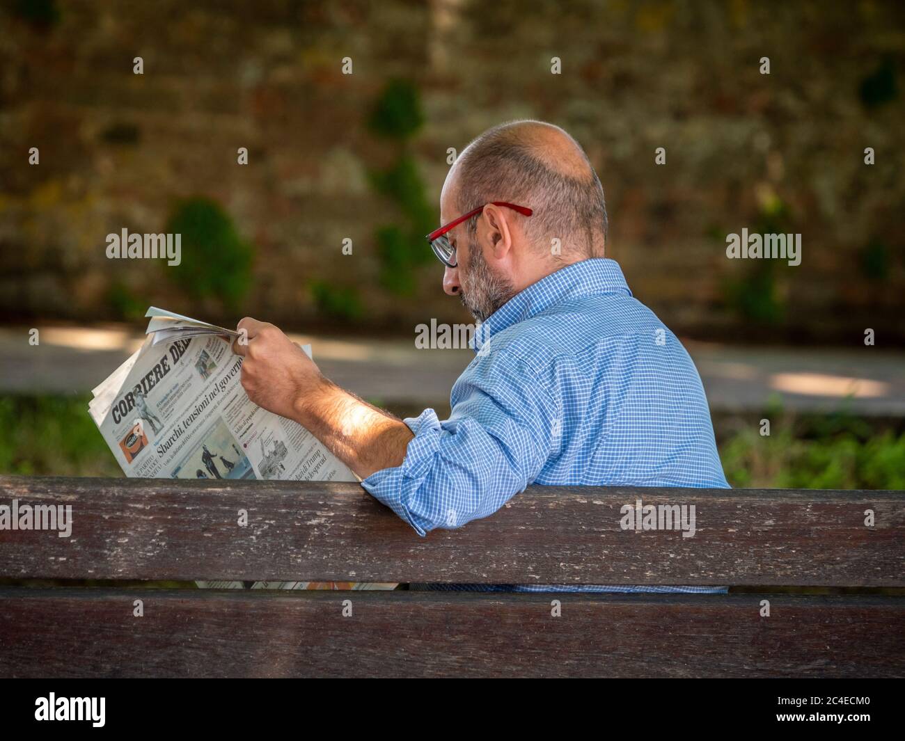 Vista posteriore di un uomo caucasico di mezza età seduto su una panchina che legge un giornale. Siena, Italia. Foto Stock
