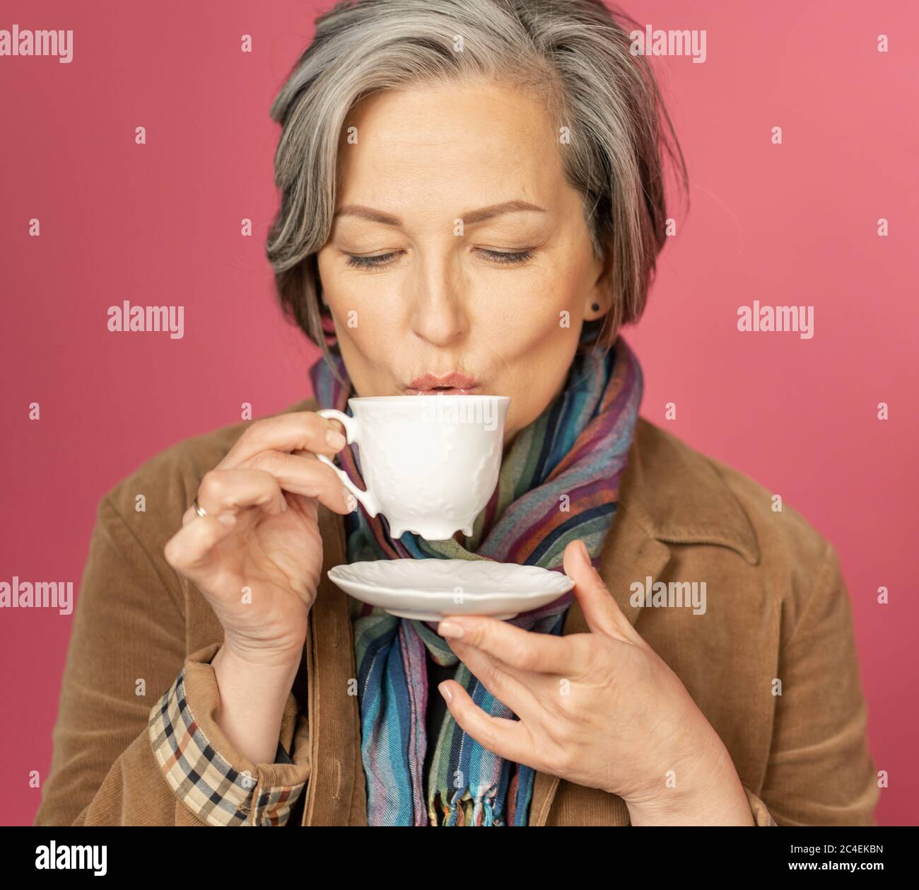 Concetto di tempo di caffè. In studio, una donna dai capelli grigi caucasici di charme beve caffè in tazza bianca su sfondo rosa. Primo piano Foto Stock