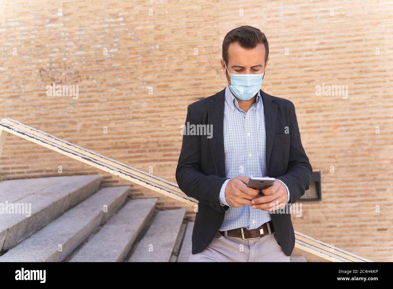 Uomo d'affari con una giacca blu e una maschera sul viso appoggiata contro una ringhiera mentre usa il suo cellulare Foto Stock