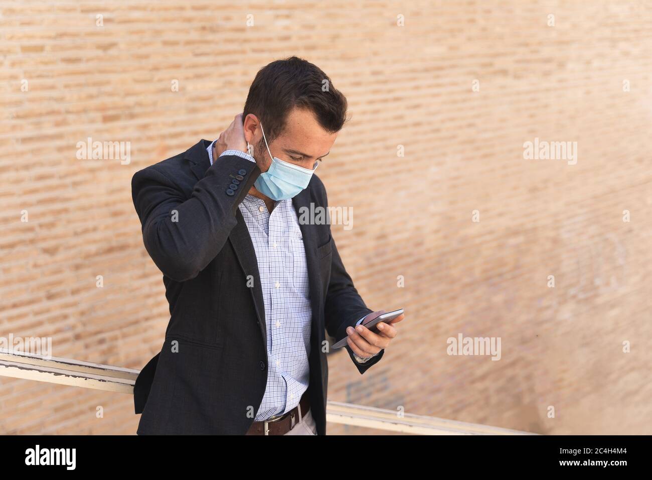 Uomo d'affari con una giacca blu e una maschera sul viso facendo un gesto preoccupato mentre si guarda il suo cellulare di fronte a un muro di mattoni Foto Stock