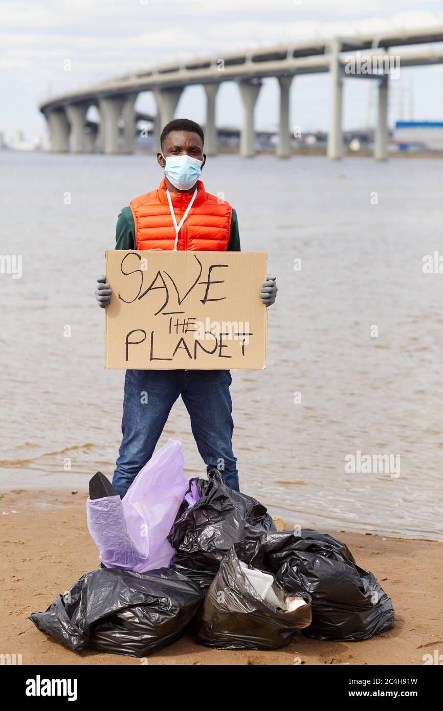 Ritratto di volontario africano in maschera protettiva che tiene il cartello mentre si trova vicino ai sacchi per rifiuti vicino al fiume Foto Stock