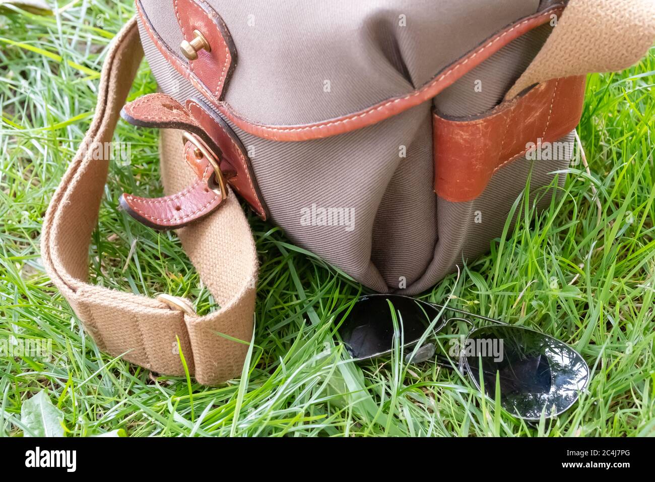 Un paio di occhiali da sole stile aviatore visto adagiato su un'erba lussureggiante accanto a una borsa per macchina fotografica in stile tela. Foto Stock