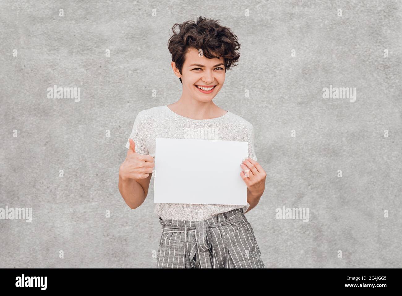 Giovane donna graziosa con i sorrisi creativi dell'acconciatura e tiene il pezzo bianco di carta nelle sue mani. Bock up. Spazio per copiare il testo. Foto Stock