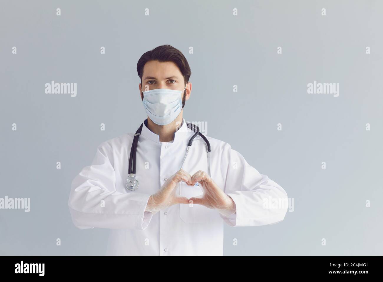 Giovane medico maschile in maschera medica che mostra il gesto del cuore su sfondo grigio. Salute cardiovascolare, carità o donazione Foto Stock