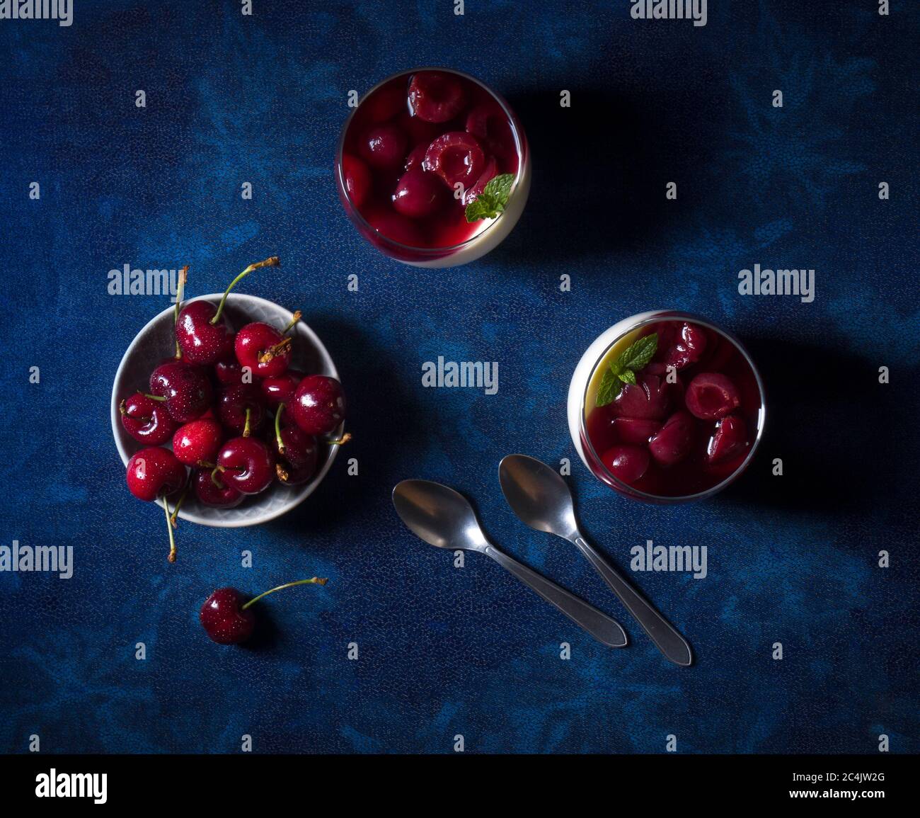 Panna cotta dessert tradizionale italiano con ciliegie in camicia al rum e sciroppo di miele. Tavolo di fondo blu scuro, ciotola delle ciliegie fresche, 2 bicchieri Foto Stock