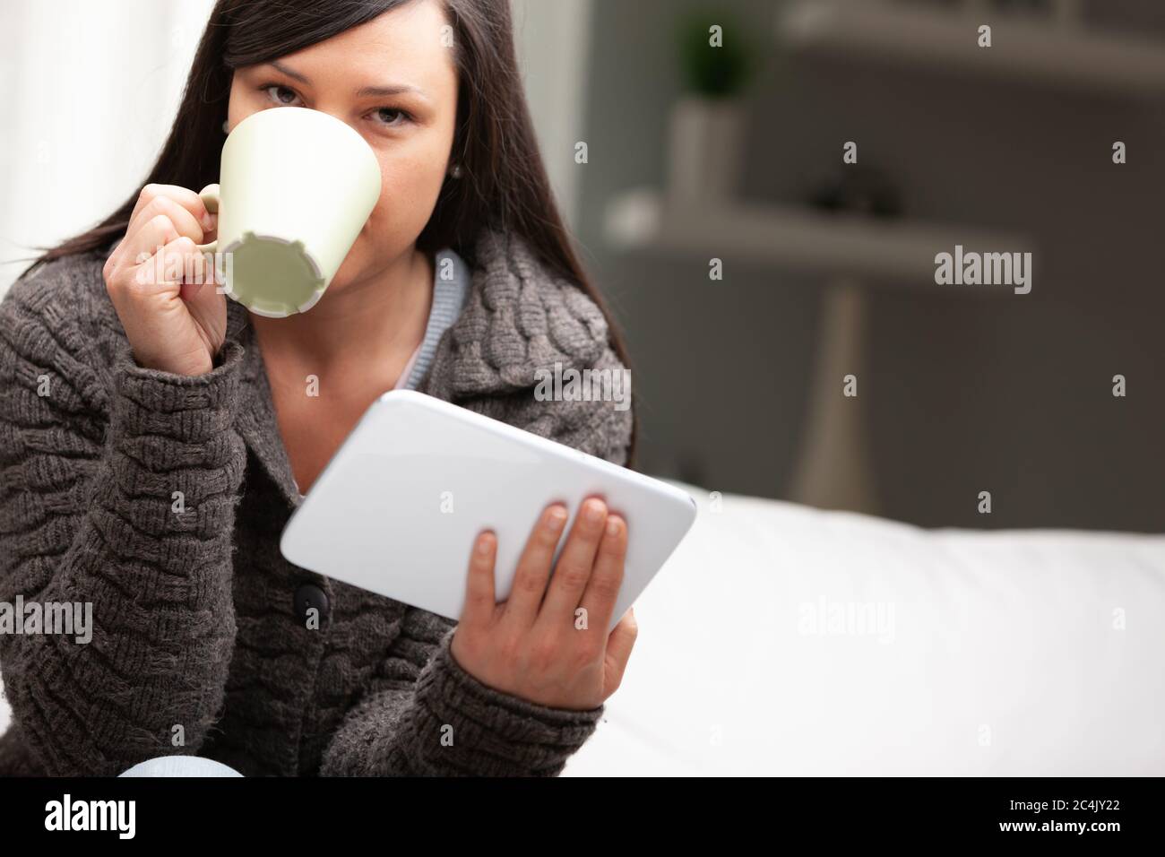 donna nel suo soggiorno che ha la colazione con la tazza di tavoletta e brioche, leggendo notizie e iniziando il suo lavoro da casa Foto Stock