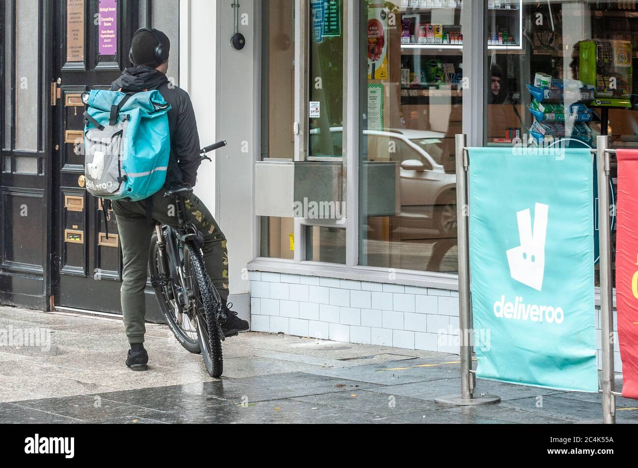 Deliveroo Food Delivery Rider in attesa di un ordine accanto a un cartello pubblicitario Deliveroo a Cork City, Irlanda. Foto Stock