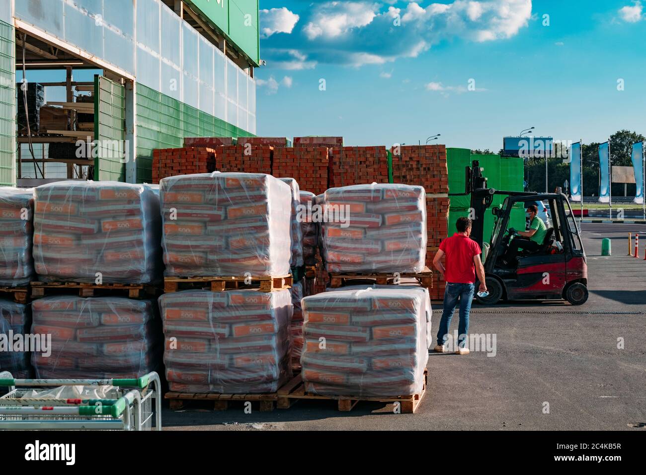 Pacchi impilati con materiali da costruzione vicino al magazzino. Il caricatore a forche è in funzione. Foto Stock