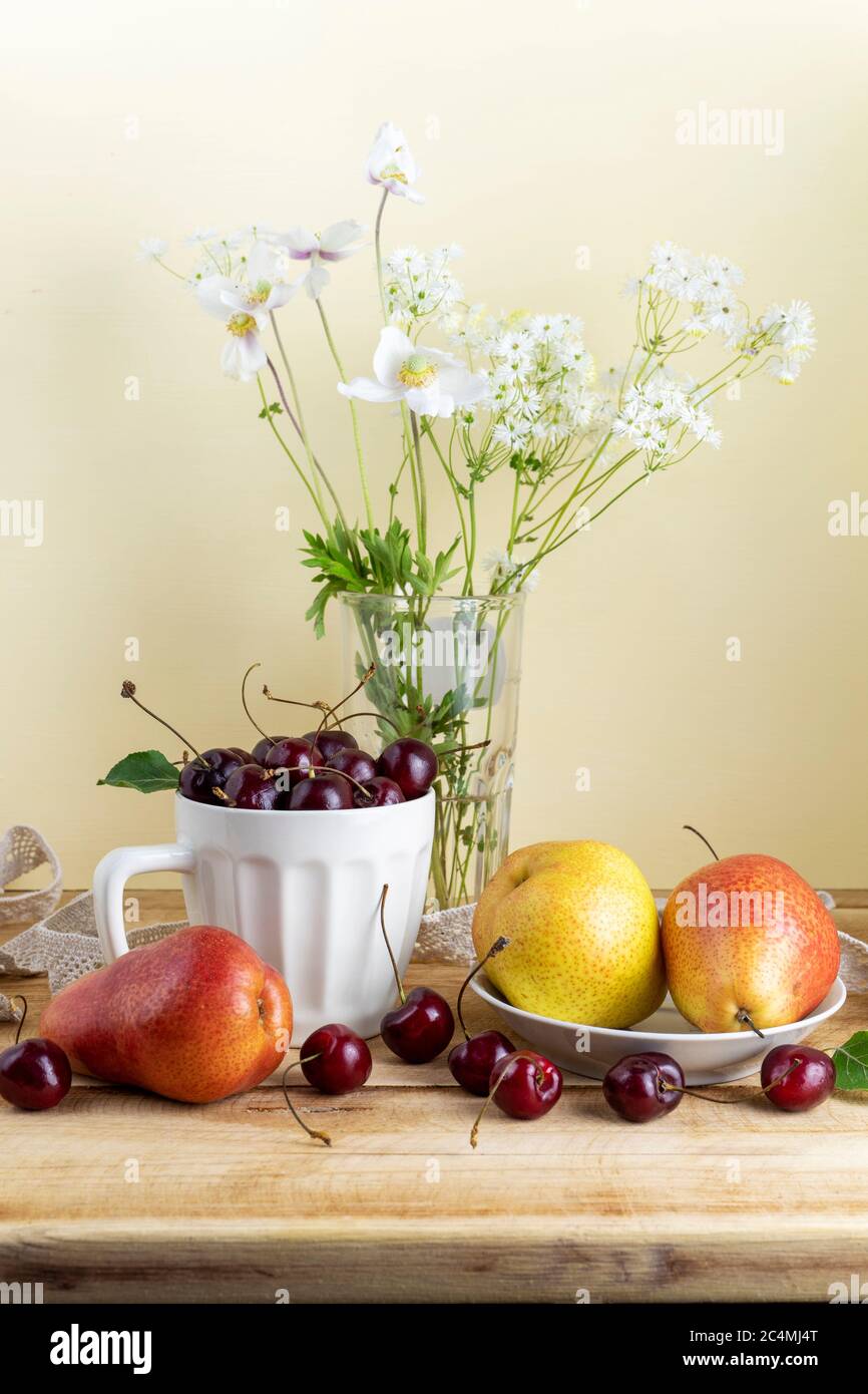 Ancora vita con pere mature, ciliegie e fiori selvatici su tavola di legno. Sfondo accogliente con illuminazione soffusa al mattino Foto Stock