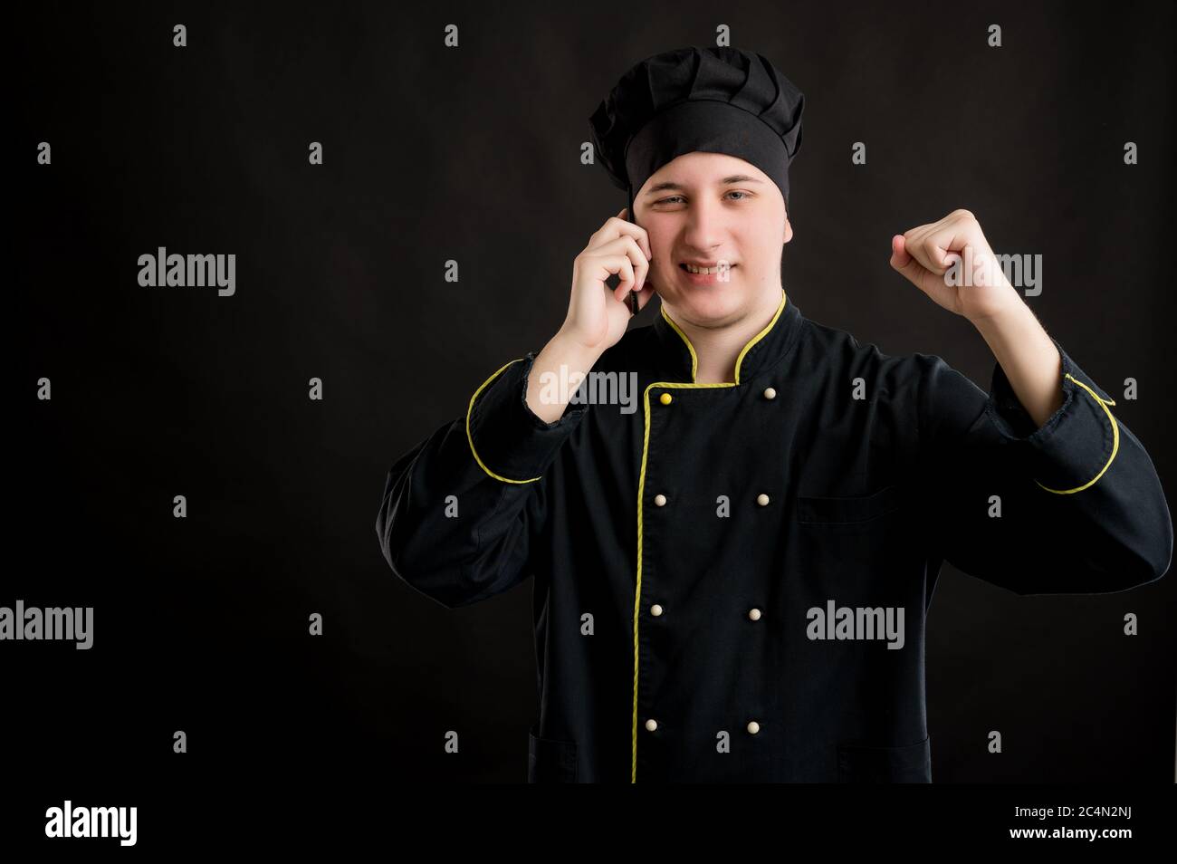 Ritratto di giovane maschio vestito da cuoco nero con, volto felice e gesti e parlando sul cellulare, in posa su uno sfondo nero isolato. Foto Stock