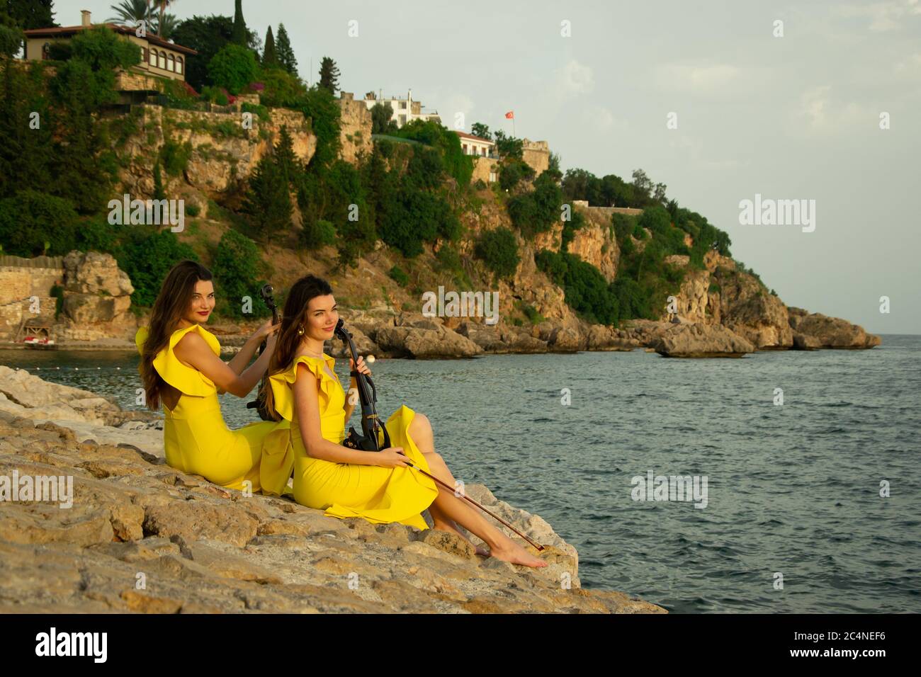 Due belle sorelle violiniste in abiti da concerto gialli sono seduti su rocce vicino al mare Mediterraneo con violini elettrici nella città vecchia di Ant Foto Stock