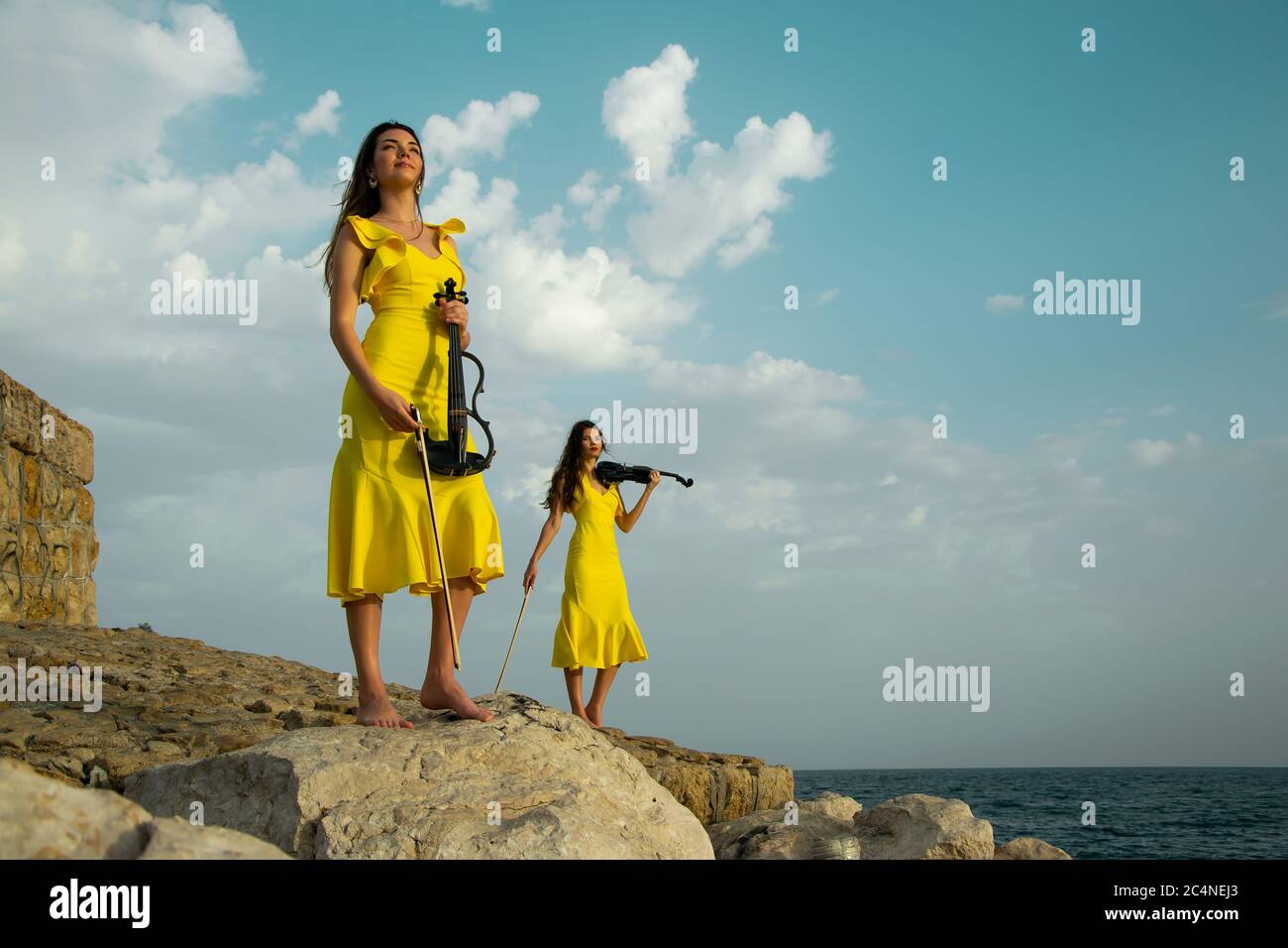 Due belle sorelle violiniste in abiti da concerto gialli suonano violini elettrici su rocce sul Mediterraneo ad Antalya, Turchia. Mare Foto Stock