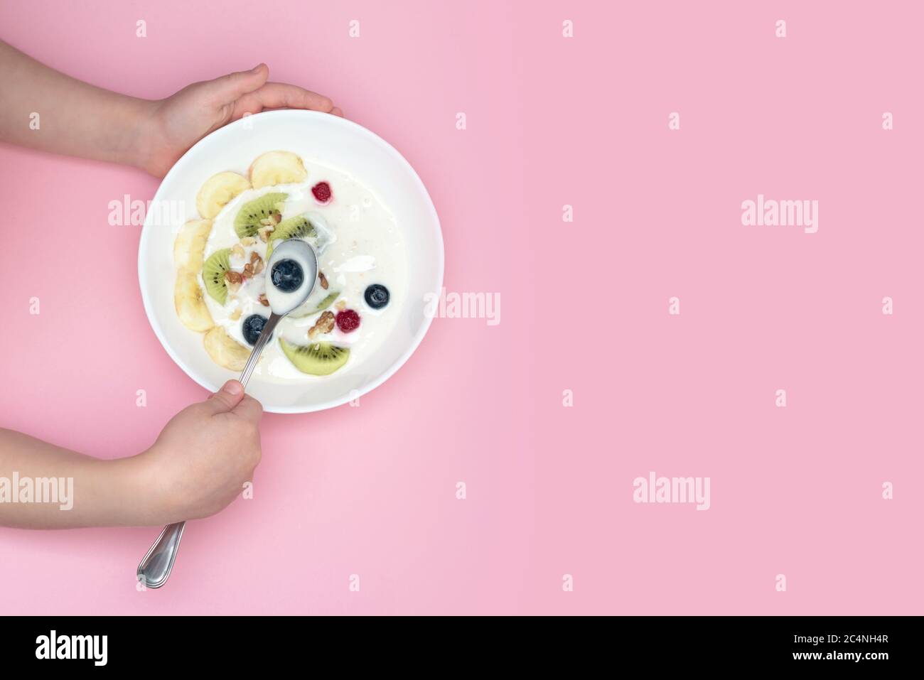 Le mani del bambino tengono una ciotola e un cucchiaio di frullato superfood. Ciotola: Yogurt, mirtilli, kiwi, banana, noci. Vista dall'alto. Foto Stock