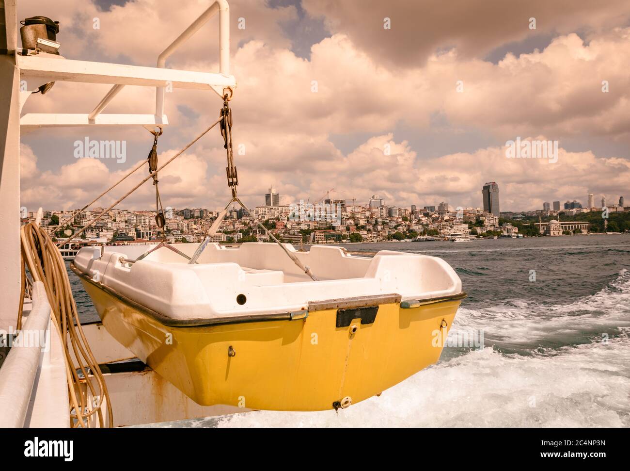piccola barca di colore giallo-bianco appesa a una nave in movimento, grandi nuvole bianche, mare schiumoso e la città dietro come vista di sfondo. Foto Stock