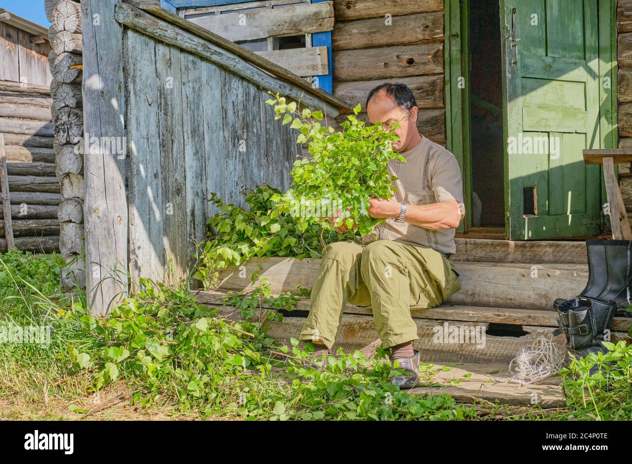 Senior Asian uomo cravatte di betulla per un bagno seduto su un portico di legno di una vecchia casa di campagna in un villaggio Siberiano, Russia Foto Stock