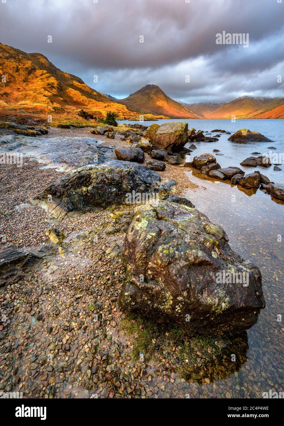 Rocce sulla costa del lago Wastwater con la bella luce dorata sulle montagne e le nuvole drammatiche scure. Lake District National Park, Regno Unito. Foto Stock