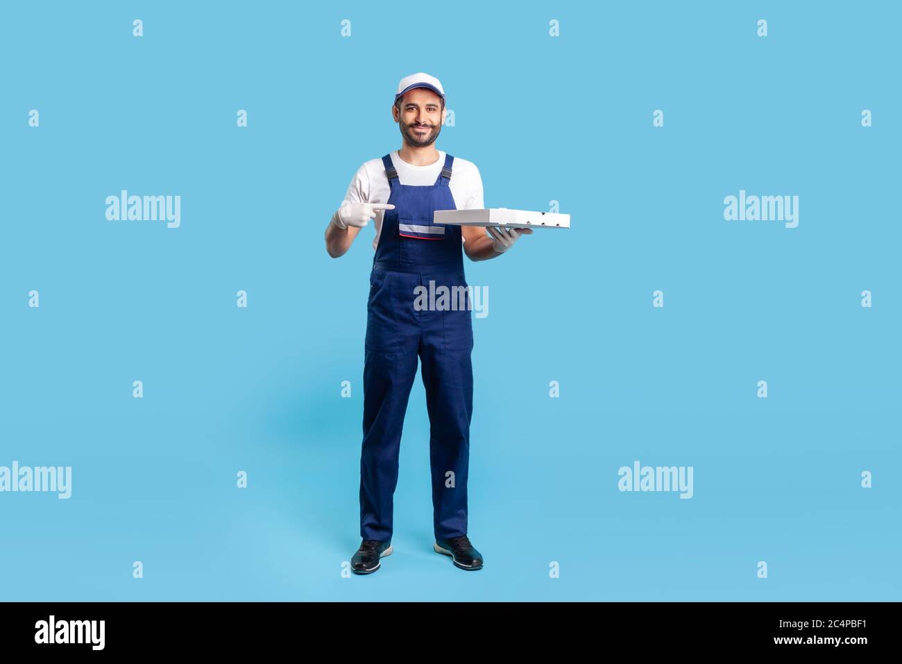 Corriere professionale a lunghezza intera in abbigliamento da lavoro e  guanti protettivi che puntano alla scatola della pizza, sorridente  amichevole alla macchina fotografica. Servizio di consegna rapida degli  alimenti Foto stock -