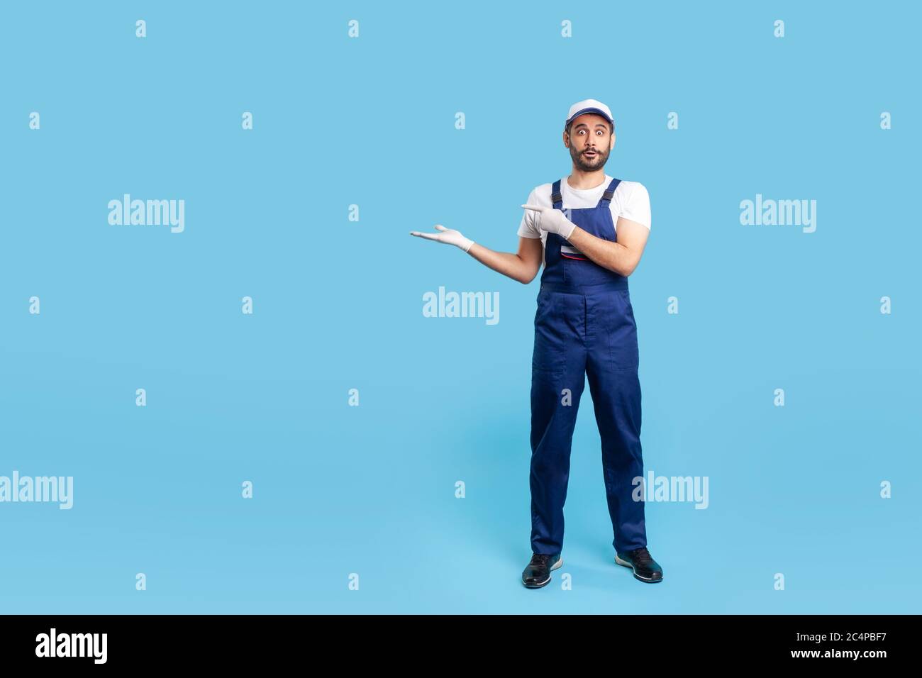 Workman sorpreso a tutta lunghezza in tute blu, cappuccio e guanti protettivi in piedi, puntando spazio vuoto sul braccio e guardando con impressionato ex Foto Stock
