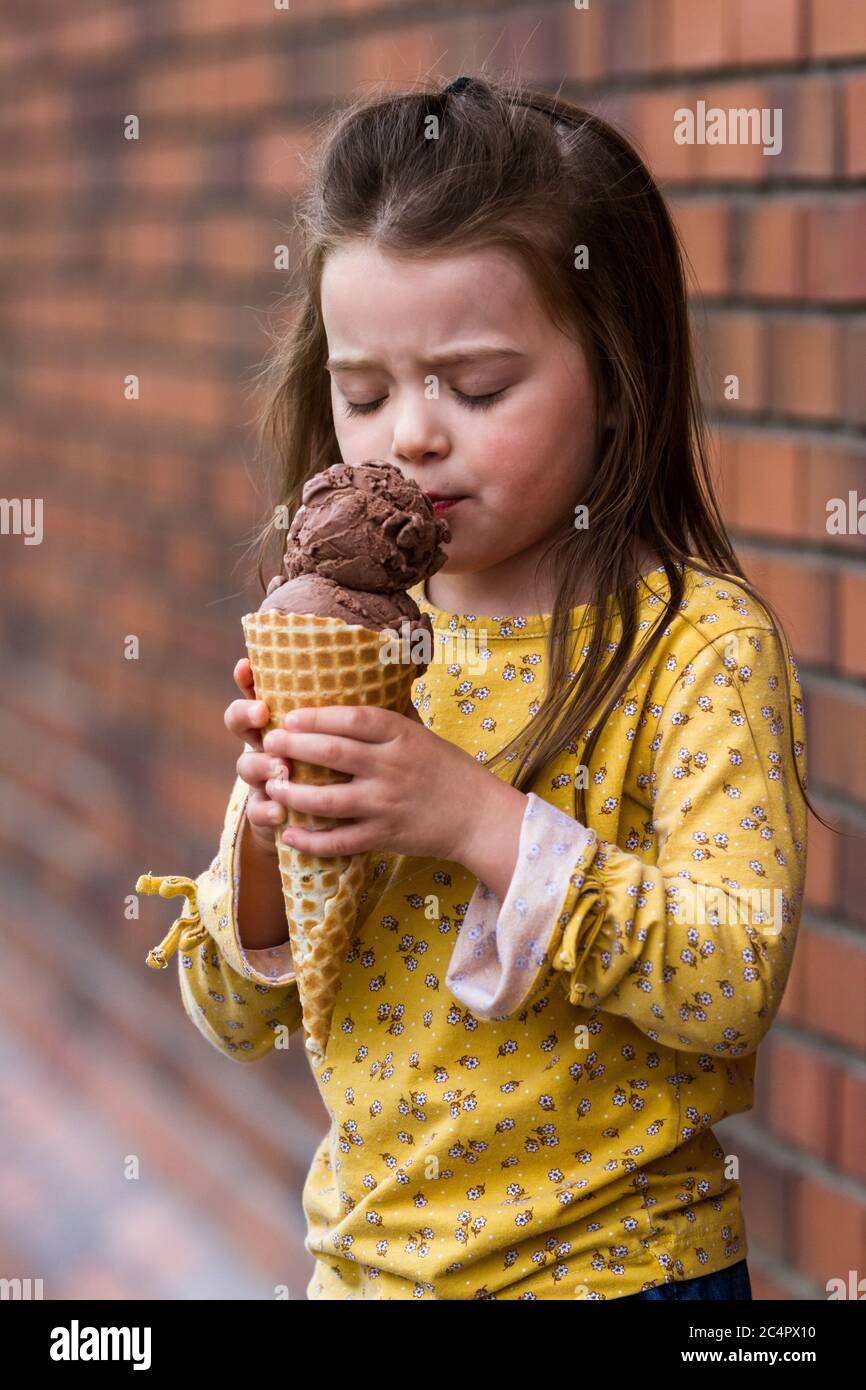 Primo piano di una adorabile bambina che mangia un grande cono gelato al cioccolato con un muro di mattoni sullo sfondo Foto Stock