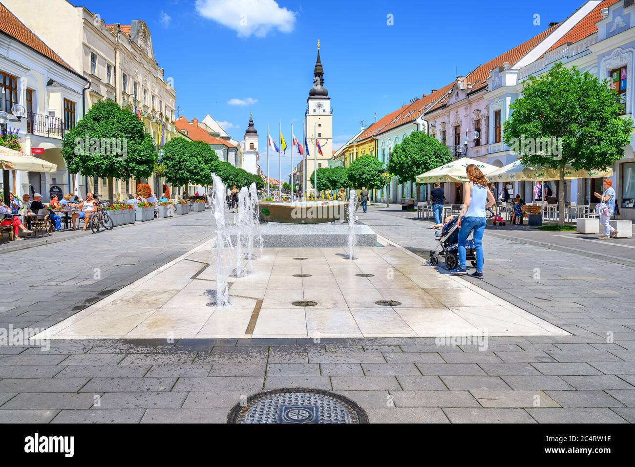 TRNAVA, SLOVACCHIA – GIUGNO 25 2020: Fontana d'acqua nella zona pedonale del centro di Trnava con torre cittadina sullo sfondo. Foto Stock