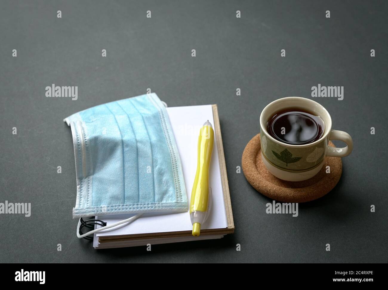 Tazza da caffè con maschera medica, blocco note e penna su rustico piano portapaziente nero. Spazio di copia. Foto Stock