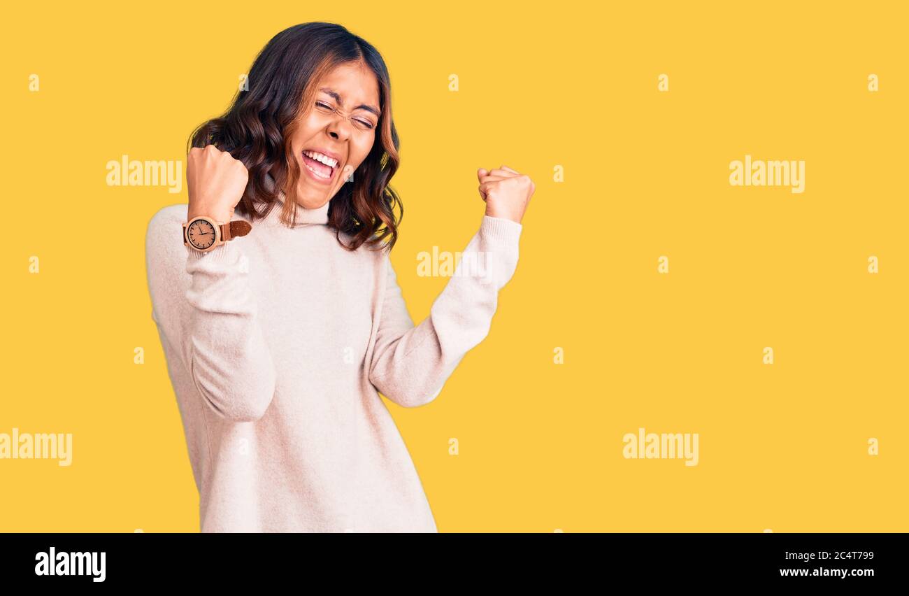Giovane bella donna mista da corsa con maglione invernale a dolcevita molto felice ed eccitata facendo gesto vincitore con braccia sollevate, sorridenti e urlanti Foto Stock