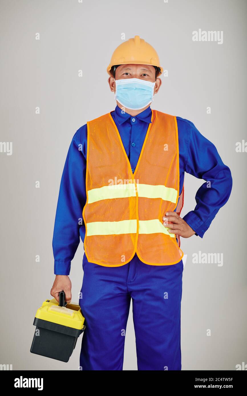 Ritratto di handyman in hardhat, gilet e maschera medica che porta cassetta attrezzi Foto Stock
