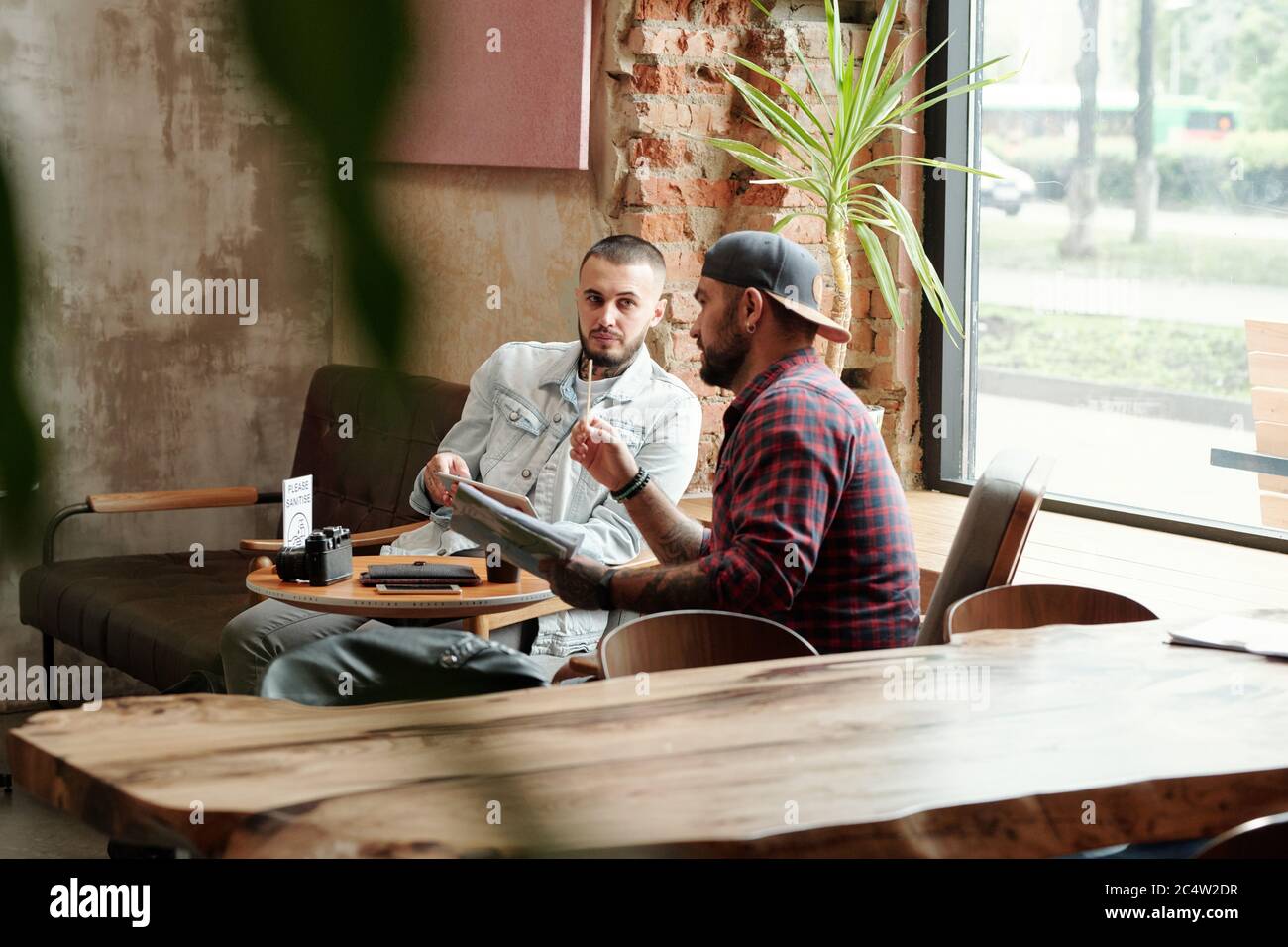 Fotografo hipster in berretto da palla seduto al tavolo al bar e discutere piano di ripresa fotografica con l'assistente dopo la quarantena del coronavirus Foto Stock