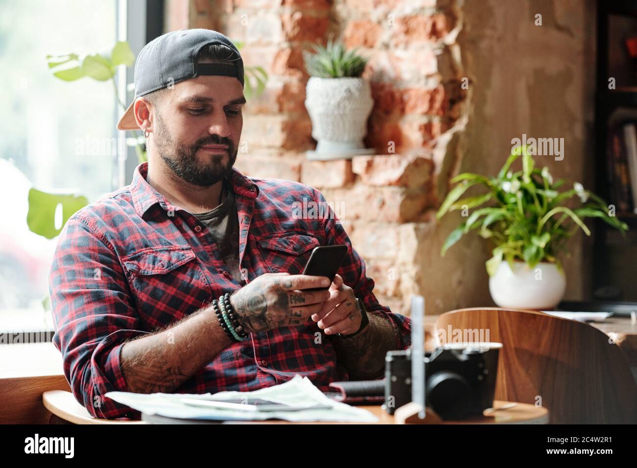 Un ragazzo brutale in berretto seduto in un accogliente bar e leggendo sms sullo smartphone mentre si ha una pausa in città Foto Stock