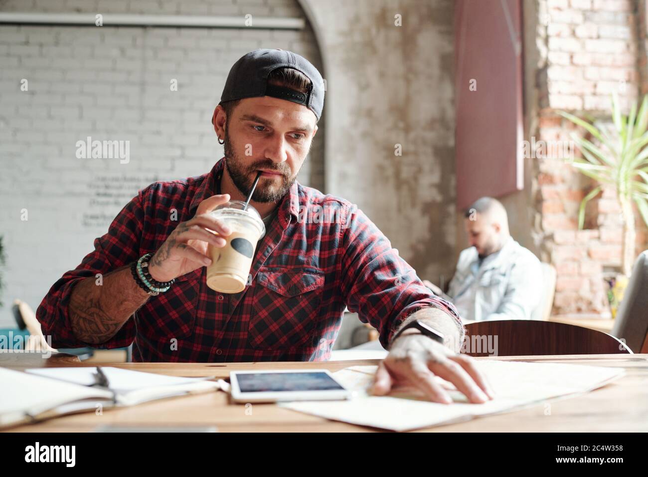Uomo giovane serio bearded in berretto di sfera che beve il caffè freddo e che controlla i posti sulla carta della mappa in caffè Foto Stock