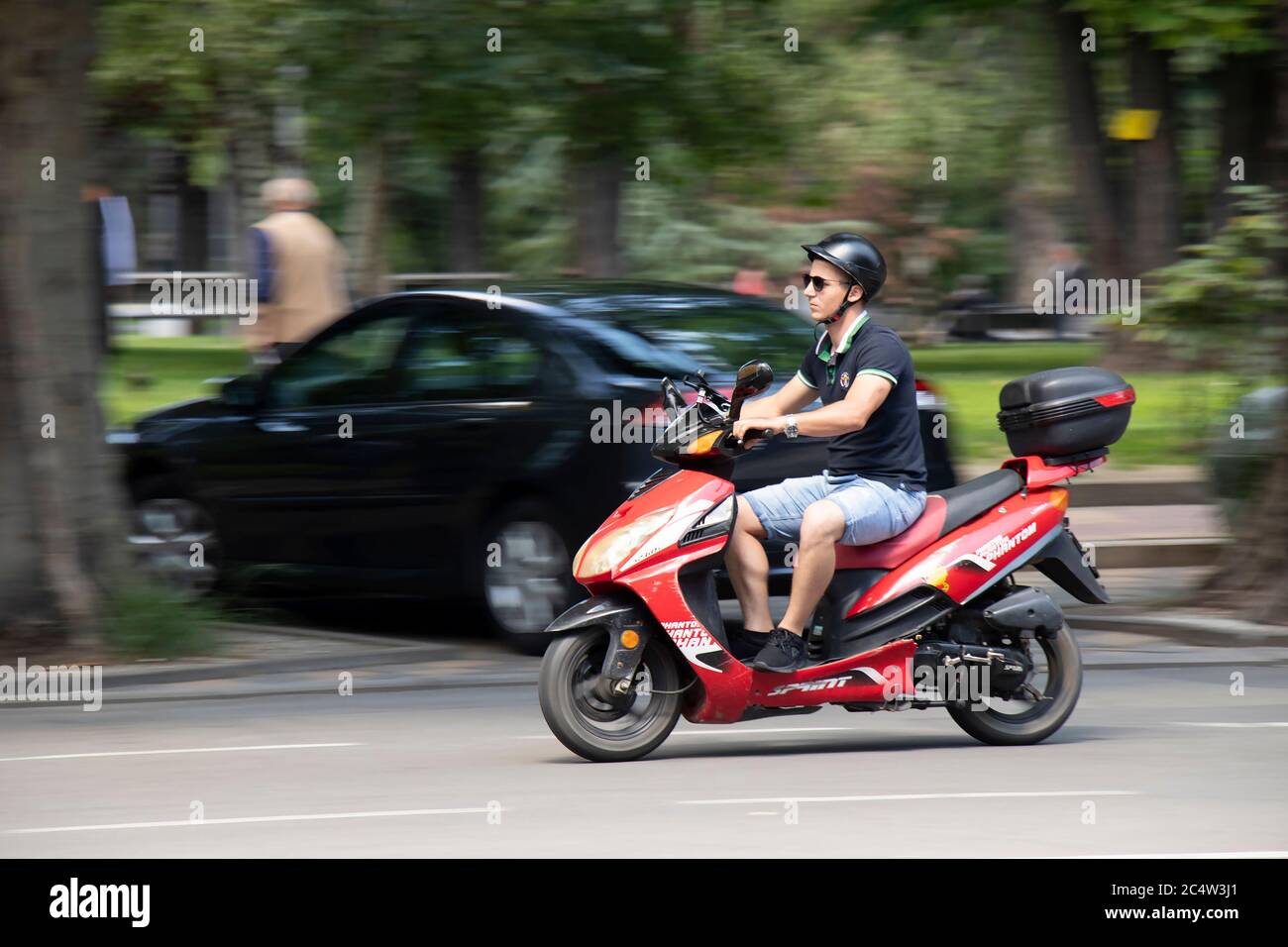 Belgrado, Serbia - 25 giugno 2020: Giovane uomo in pantaloncini e t-shirt in sella a uno scooter rosso con box posteriore sulla strada della città, vista laterale Foto Stock