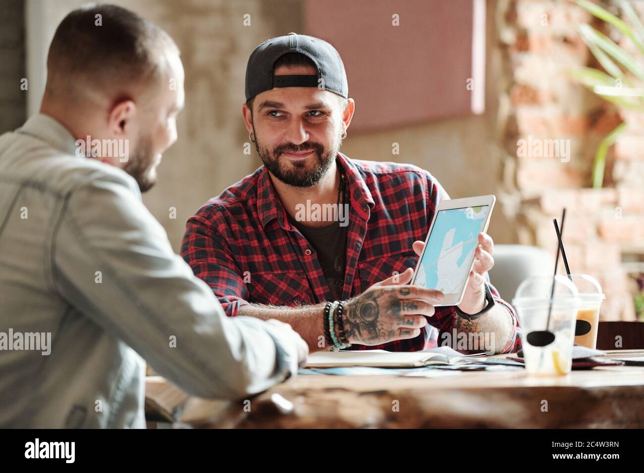 Contenuto bell'uomo in berretto che mostra il percorso di viaggio verso un amico mentre pianificano il viaggio in bar Foto Stock