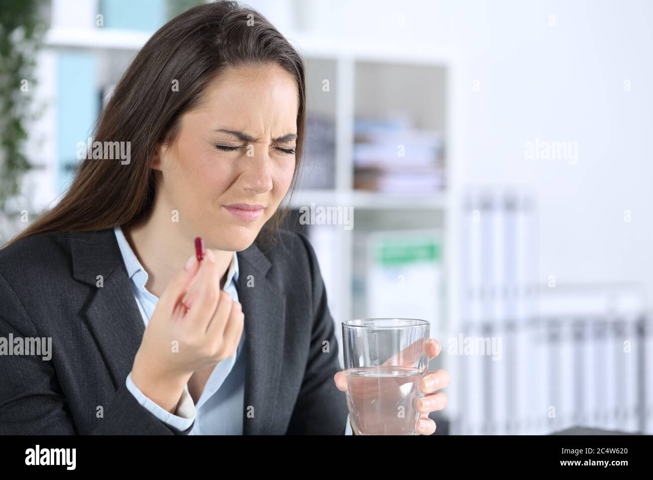 Donna dirigente malata che tiene la pillola antidolorororororororante e il bicchiere d'acqua seduto su una scrivania in ufficio Foto Stock