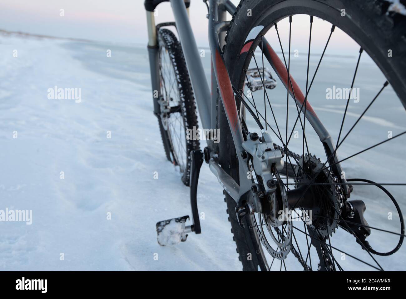 Mountain bike soggiorno nella neve vicino al mare ghiacciato o lago. Dettaglio del meccanismo di cambio marcia e delle rotture del disco. Sfruttamento e manutenzione delle biciclette invernali. Con borchie Foto Stock