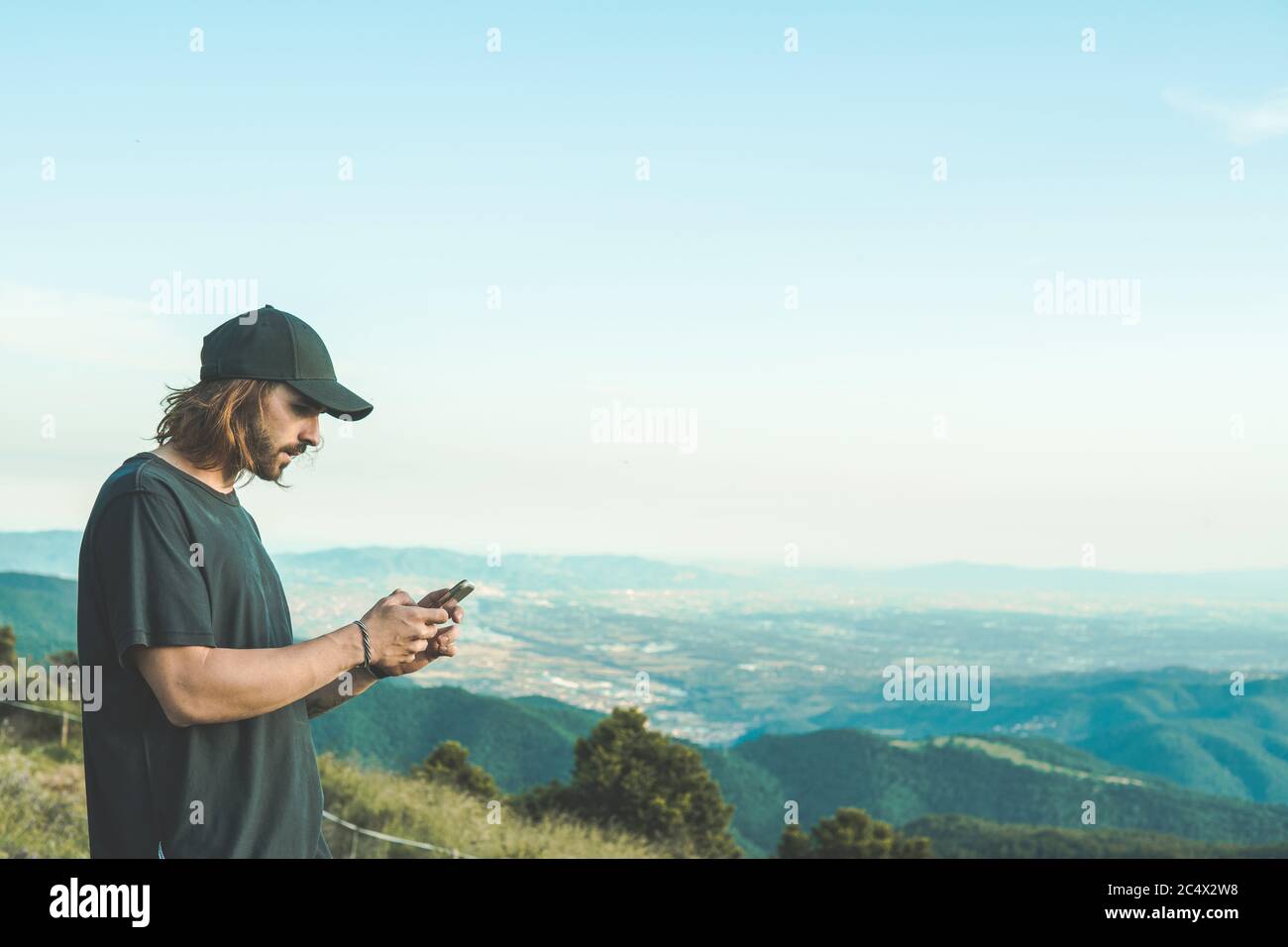 Bel giovane con un cappuccio e capelli lunghi. Sta navigando in Internet sul suo telefono cellulare. La vista sulla valle è incredibile. Foto Stock