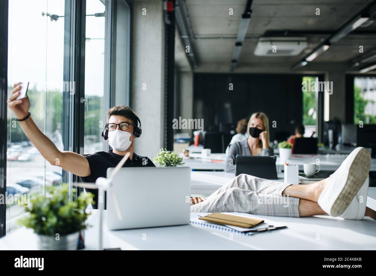 Giovane uomo con maschera di protezione al lavoro o a scuola in ufficio dopo il blocco, prendendo selfie. Foto Stock