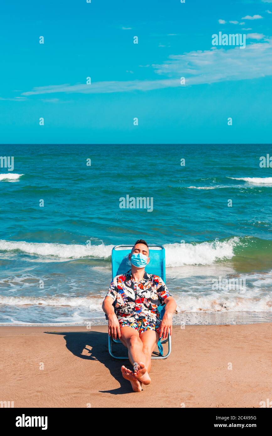 un uomo caucasico che indossa una maschera chirurgica blu mentre è seduto su una sedia a sdraio sulla spiaggia, con il mare sullo sfondo Foto Stock