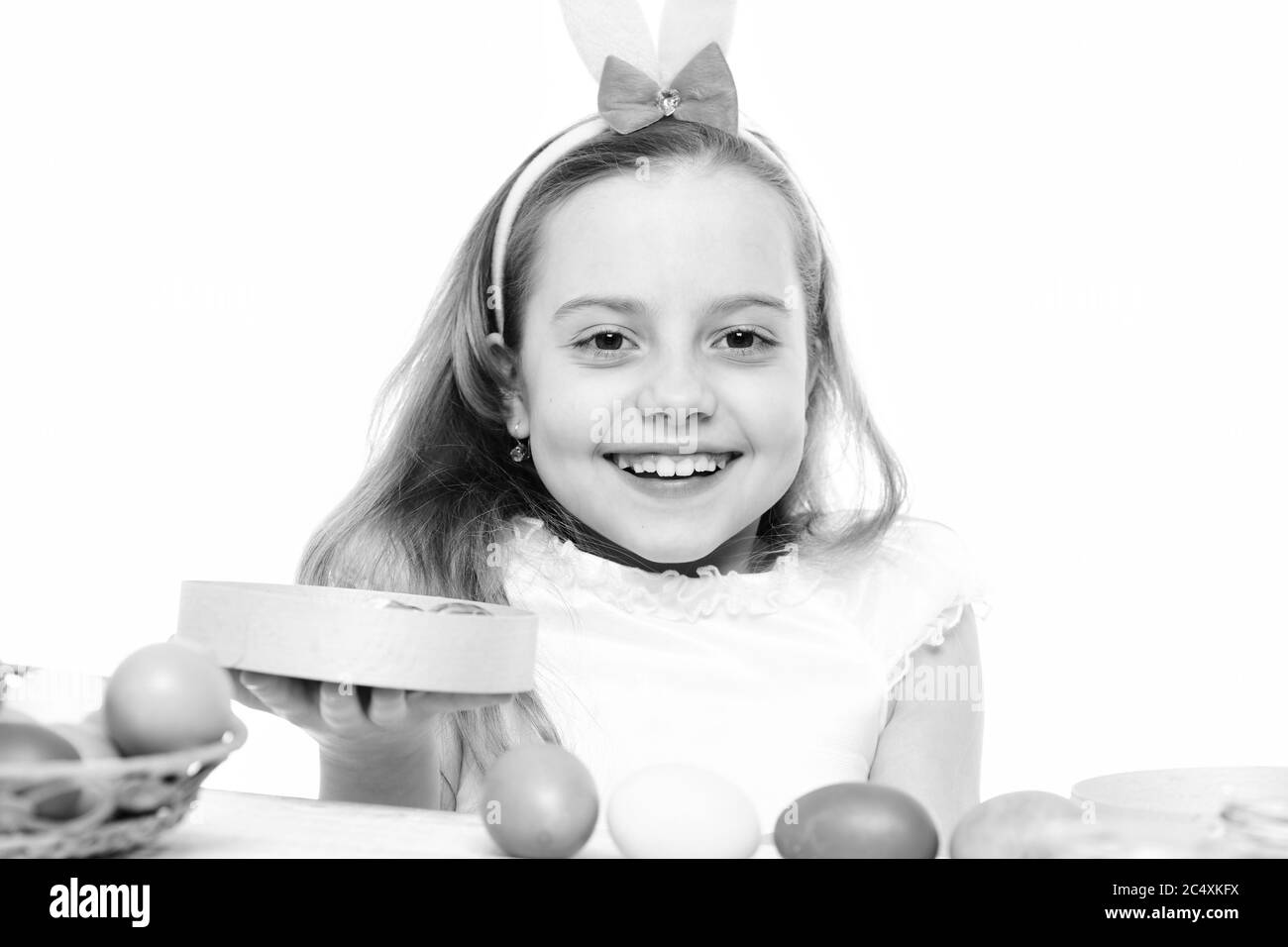 bambina piccola o bambino carino con faccia felice e orecchie rosa coniglio sulla testa bionda intorno uova di pasqua colorate tenendo scatola isolato su sfondo bianco. Foto Stock