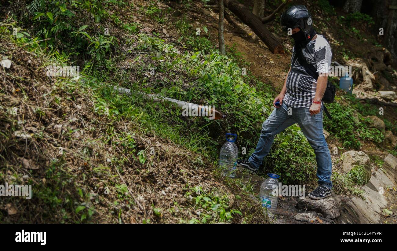 Caracas, Venezuela. 28 Giugno 2020. Un uomo che indossa un casco e una protezione per la bocca riempie un canestro di acqua in corrispondenza di un tubo sciolto nella parte est della capitale. Nel mezzo della pandemia di Corona, i residenti in molti luoghi del Venezuela si lamentano della mancanza di acqua. Credit: Rafael Hernandez/dpa/Alamy Live News Foto Stock