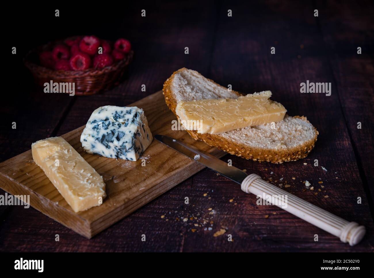 Pane con cheddar stagionato e formaggio blu morbido su tavola di legno con cesto di lamponi su sfondo scuro. Foto Stock