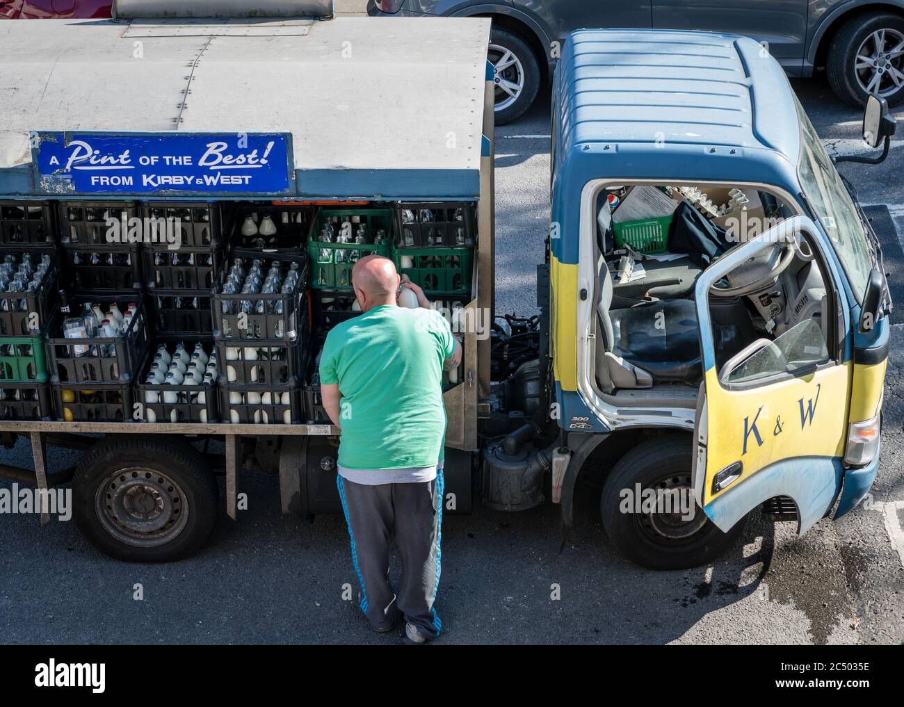 Un mungitore che effettua una consegna prendendo bottiglie di latte dal suo veicolo elettrico e consegnando direttamente alla porta del cliente. Un'azienda che sta morendo fuori. Foto Stock