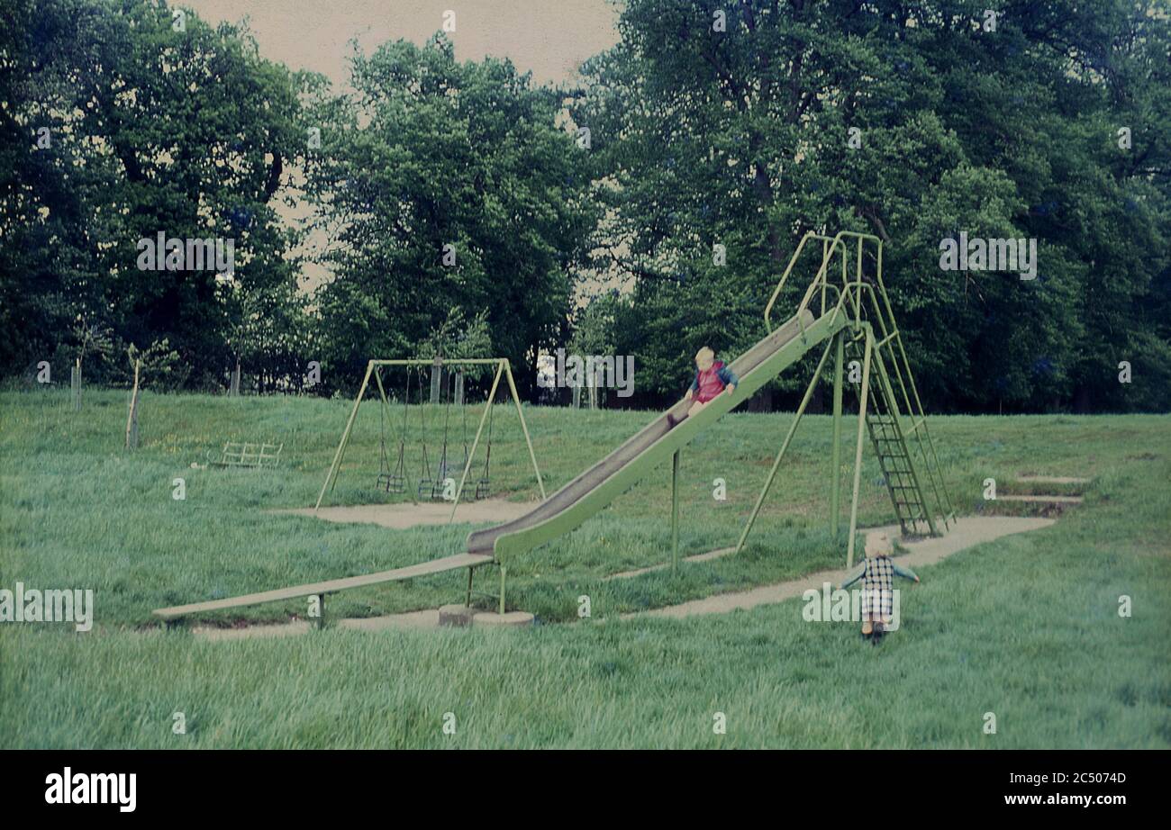 1967, due bambini piccoli che giocano all'esterno in un parco pubblico o in un campo ricreativo, con un bambino su uno scivolo in metallo tradizionale. L'attrezzatura da gioco costruita in acciaio vecchio stile, spesso risalente al periodo vittoriano, era una vista comune in parchi in tutta la Gran Bretagna in questa epoca, spesso situato come visto qui, posizionato casualmente in aree erbose senza troppo pianificazione. Foto Stock