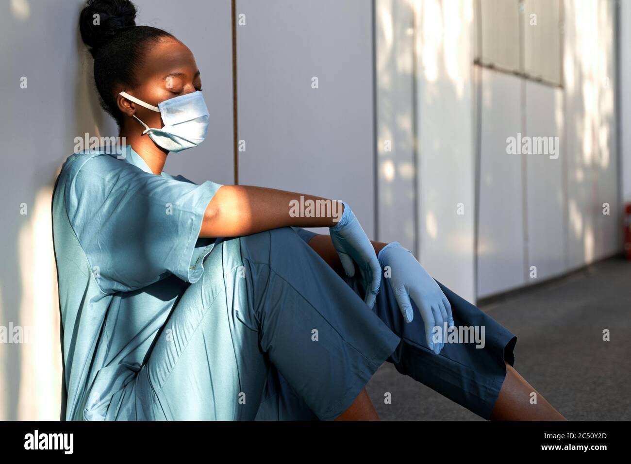 Stanco donna esausta infermiera africana indossare guanti maschera seduta sul pavimento dell'ospedale. Foto Stock