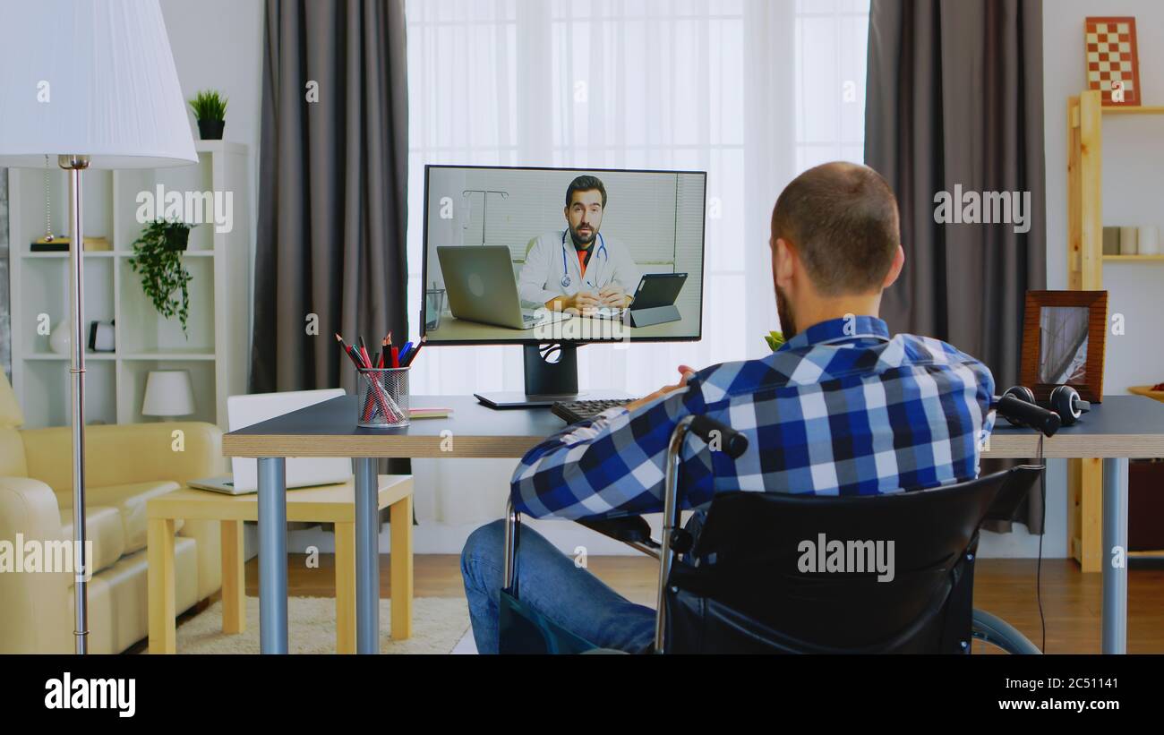Uomo con disabilità motoria in sedia a rotelle che parla con il medico in videochiamata. Foto Stock