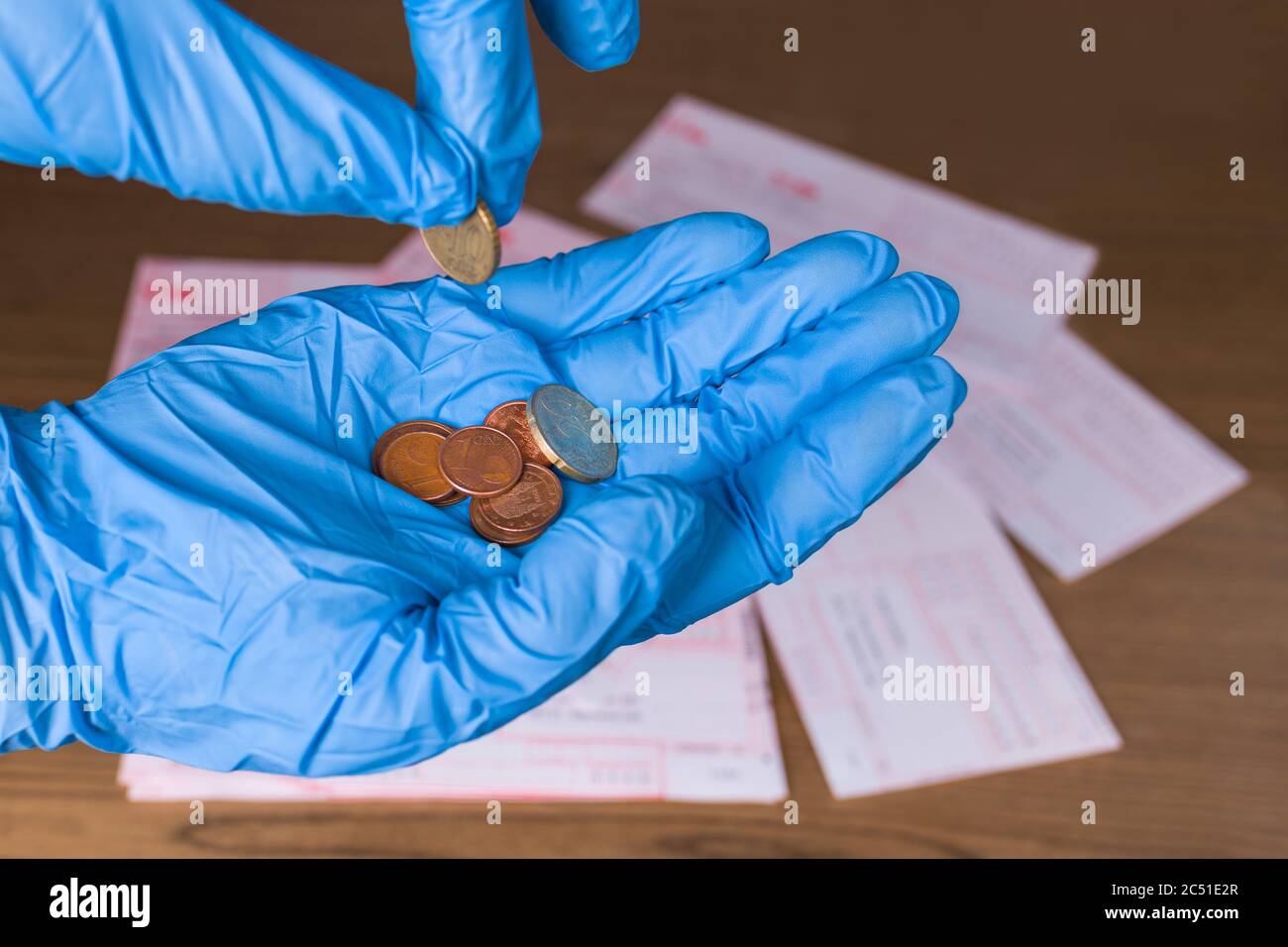 Monete metalliche in mano umana e ordini di denaro su scrivania in legno. Contando euro in contanti sul palmo in guanto protettivo blu. Pagamento delle fatture in pandemia COVID-19. Foto Stock