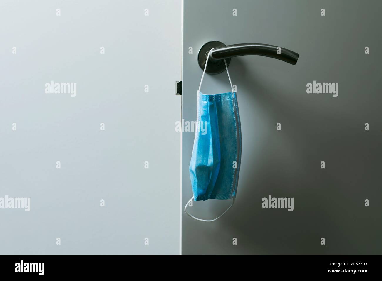 chiusura di una maschera chirurgica blu appesa al moderno pomello di una porta bianca aperta Foto Stock