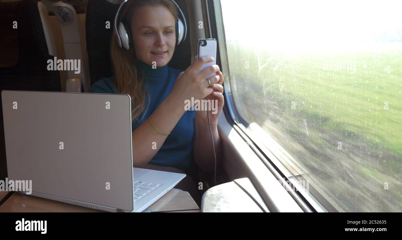 Donna che viaggia in treno con dispositivi moderni. Ascolta la musica con le cuffie wireless, scattando foto della vista esterna con lo smartphone e Havin Foto Stock