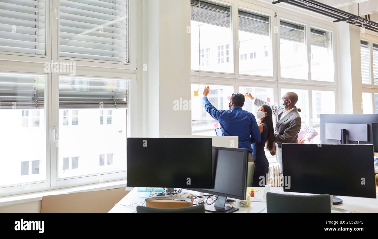 Team aziendale con maschera facciale durante il brainstorming creativo con note appiccicose sulla finestra in ufficio Foto Stock