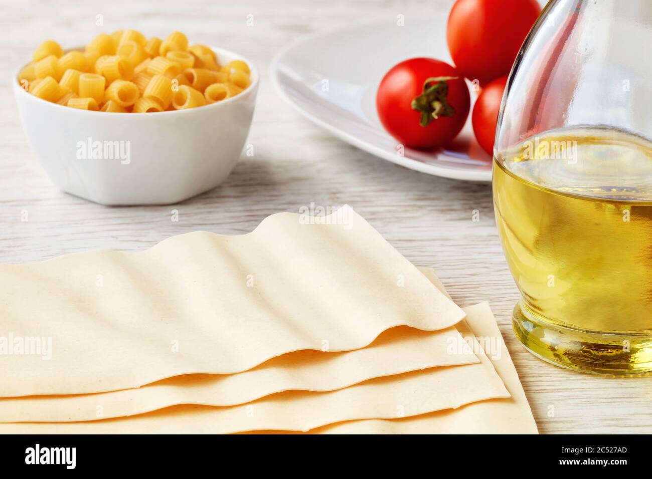 Fogli di pasta secca di lasagne non cotte con olio d'oliva e pomodori ciliegini su tavolo da cucina in legno bianco. Foto Stock