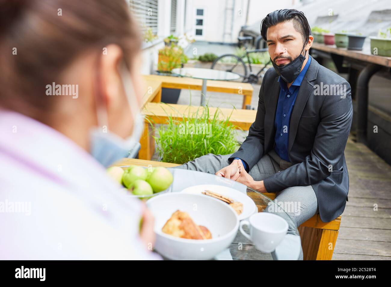 Uomo d'affari con maschera sul mento siede durante la pausa pranzo in un bar con un collega Foto Stock